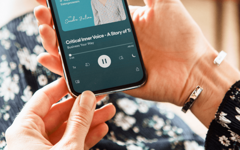 A women wearing a black and white floral dress holds a phone. On the screen is a preview of Holly's interview on the 'Business Your Way' podcast, where Holly spoke about overcoming your critical inner voice