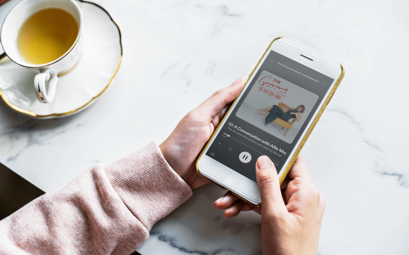 A women wearing a comfy pink sweater holds an iphone over a marble tabletop, next to a while and gold cup and saucer containing herbal tea. On the phone is a preview of Holly's interview on The Success Revolution Podcast, where she talks about owning and expressing your brilliance
